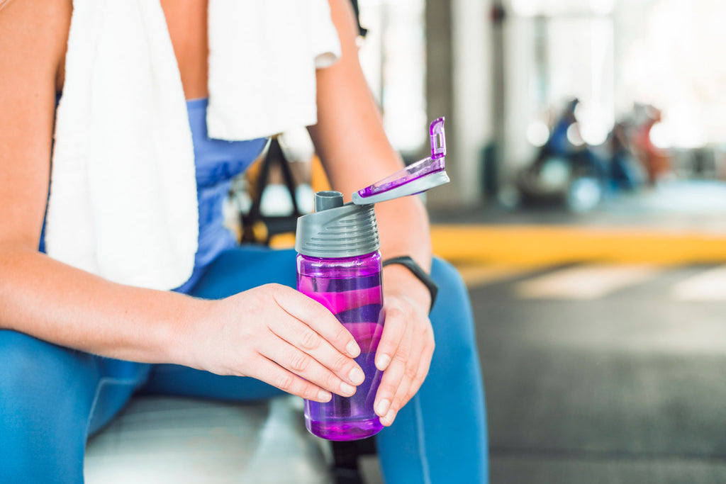 Pessoa sentada na academia, usando roupa esportiva azul e segurando uma garrafa de água roxa com tampa aberta; uma toalha branca está sobre seus ombros.
