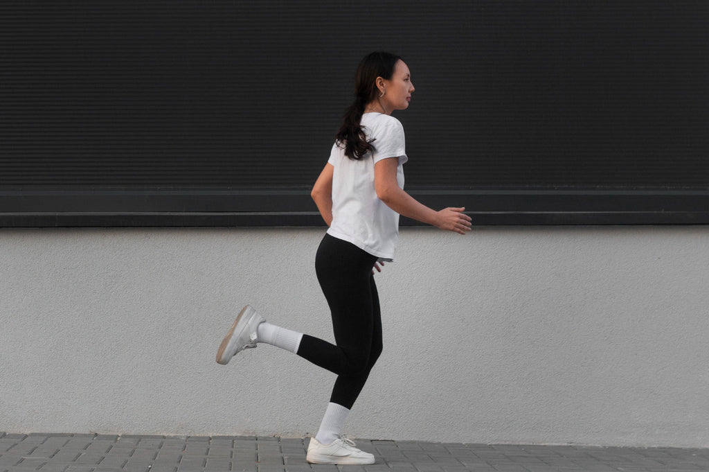 Mulher jovem praticando corrida estacionária em ambiente urbano, usando camiseta branca, legging preta e tênis esportivo.