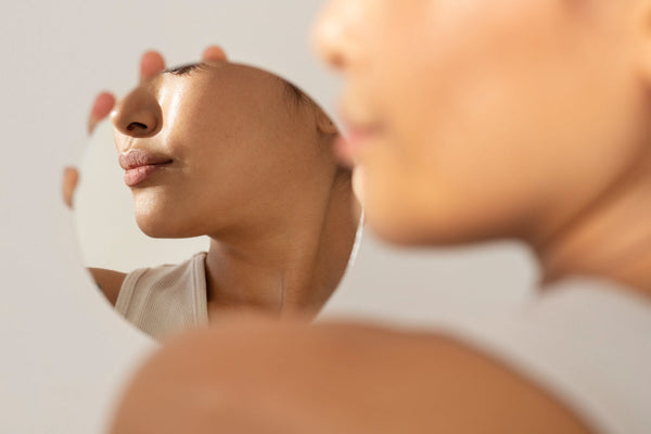 Mulher segurando um espelho redondo que reflete parte de seu rosto, com destaque para os lábios e o queixo; imagem em tons suaves e iluminação natural.