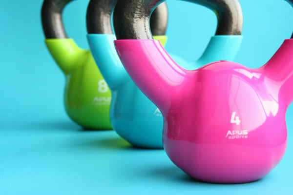 Kettlebells coloridos organizados em linha sobre fundo azul, com destaque para o peso rosa de 4 kg em primeiro plano.