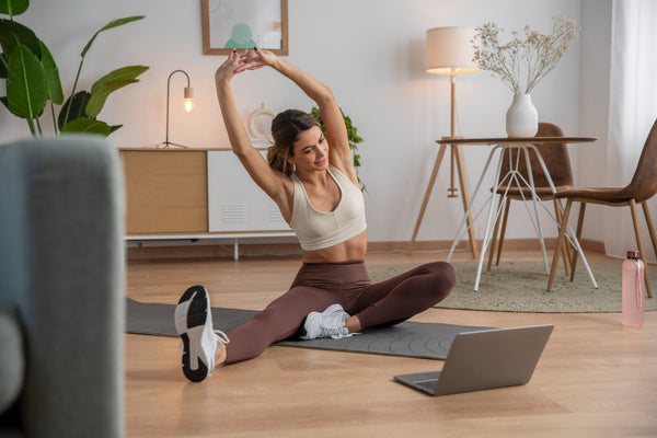 Mulher praticando alongamento em casa sobre um tapete de yoga, acompanhando treino pelo notebook; ambiente aconchegante com móveis de madeira e decoração minimalista.